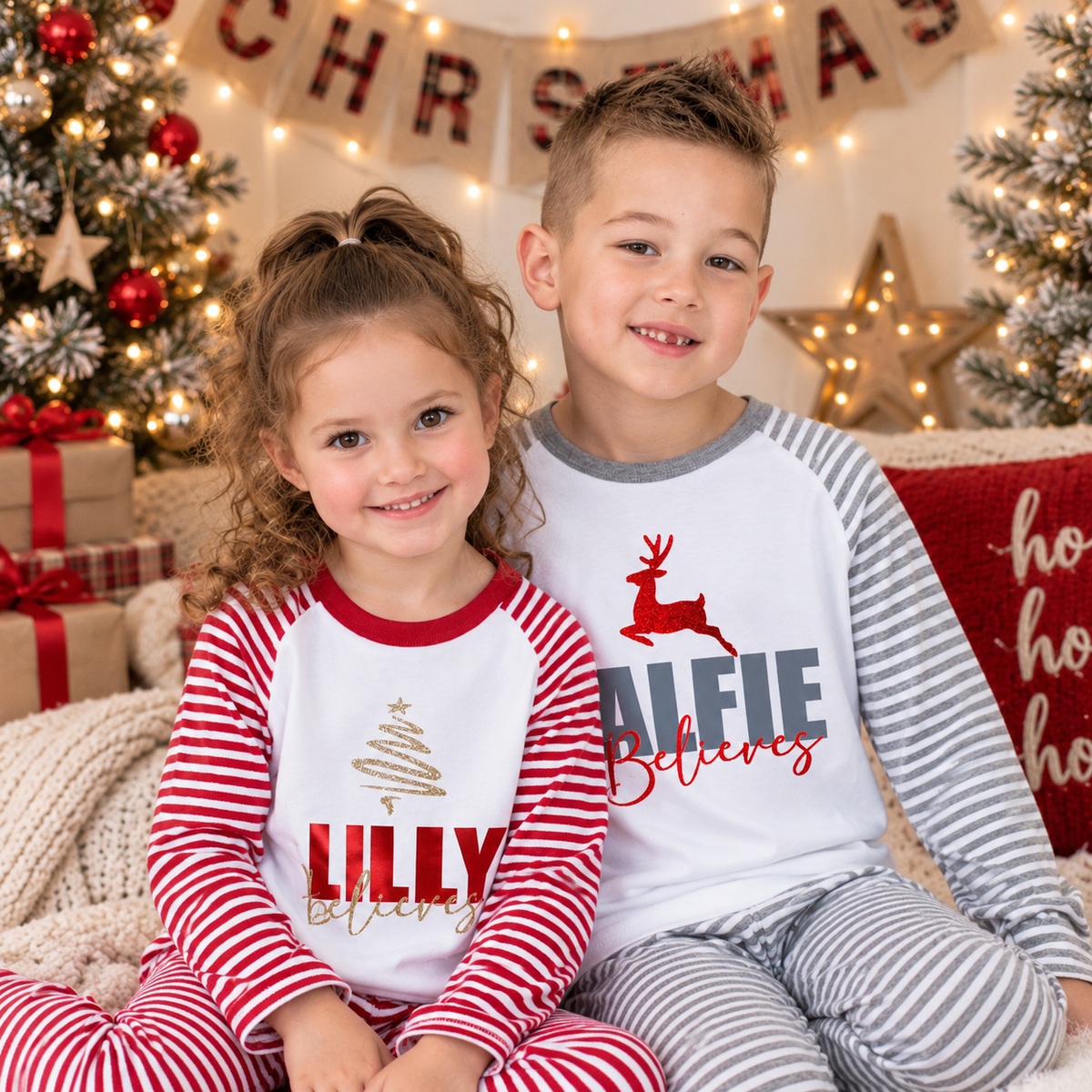 Two children wearing personalised red and grey stripe Christmas pyjamas with bold-name “Believes” designs in a festive setting by Grace & Grayson