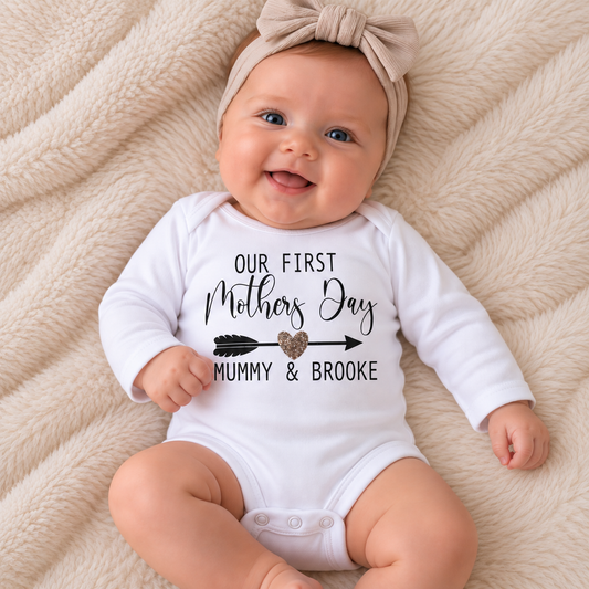 Baby wearing a white personalised Our First Mother’s Day baby vest with black arrow and gold heart design, Grace & Grayson