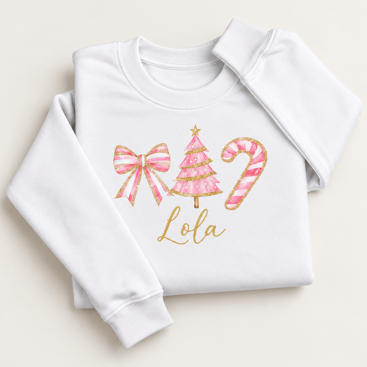 White personalised Christmas sweatshirt with a pink bow, Christmas tree and candy cane, gold detailing and script name, folded on a cream background | Grace & Grayson